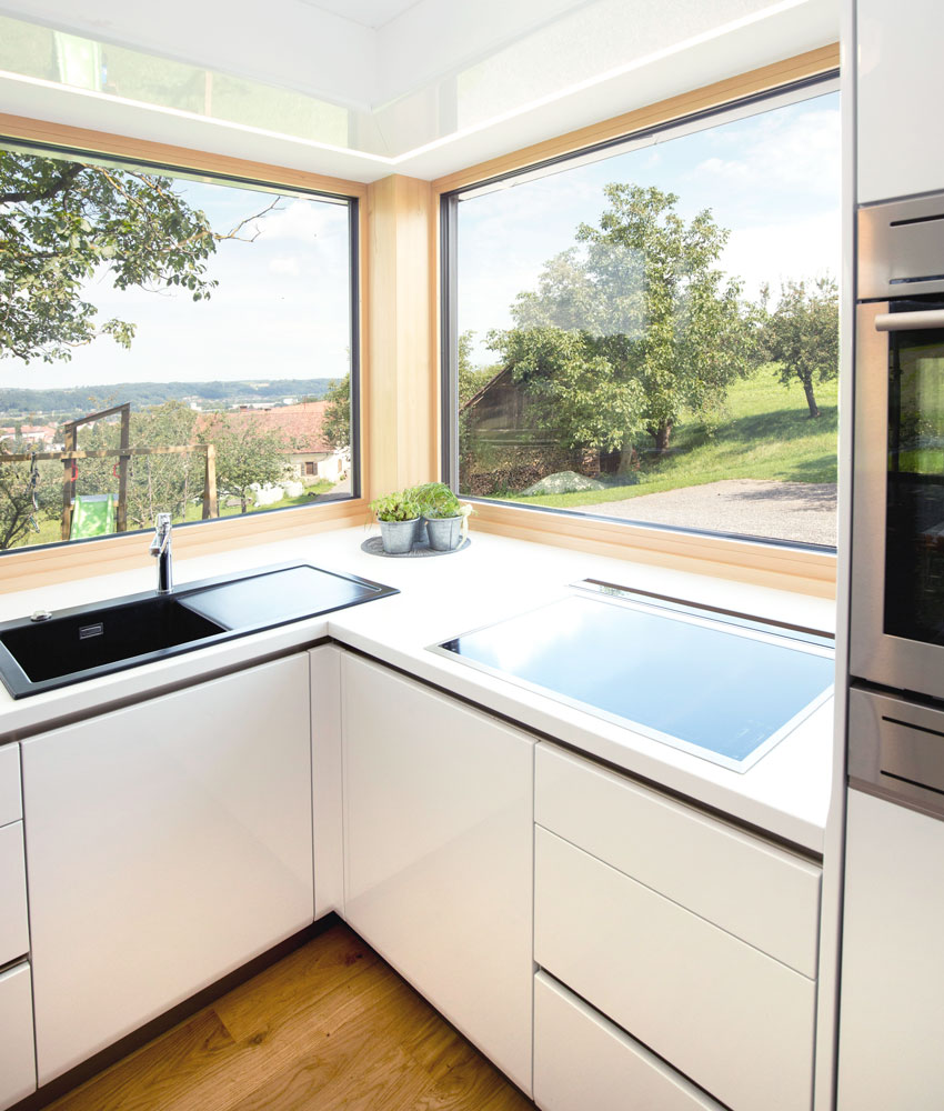 Eine maßangefertigte Küche von Tischlerei Raidl. Weiß lackierte Holzfront und weiße Oberfläche. Schwarzes Waschbecken. Ein Ceranfeld zum Kochen und ein Backofen ist ebenfalls vorhanden. Die zwei Fenster im Eck der Küche sorgen für ausreichend Beleuchtung und zeigen eine grüne Wiese mit ein paar Bäumen und einem Schuppen.